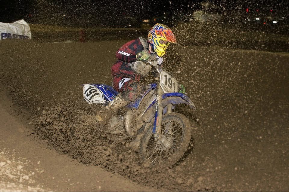 Jake racing a blue dirt bike, number 191, at night, throwing a wall of roost through a turn